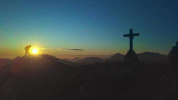 Man reaches the top of a mountain with cross at sunset