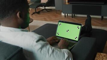 Man sitting on couch typing on laptop with green screen video