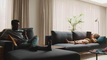 Man sitting on daybed watching smartphone and girl lying on sofa video