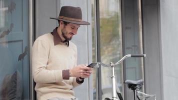 Hombre joven con estilo en el sombrero usa el teléfono celular al aire libre, hipster con bicicleta video