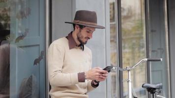 jonge stijlvolle man in hoed gebruikt mobiele telefoon buitenshuis, hipster met fiets