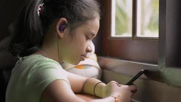 fille portant des écouteurs assis près de la fenêtre et utilisant un smartphone. video