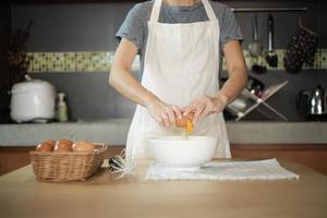 Female cook in a white apron is cracking an egg in home's kitchen. photo