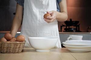Female cook in a white apron is cracking an egg in home's kitchen. photo