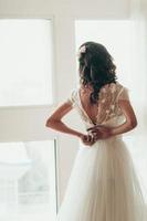 La novia abrochando su vestido junto a la ventana, vista desde la espalda foto