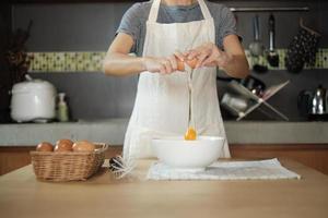 Female cook in a white apron is cracking an egg in home's kitchen. photo