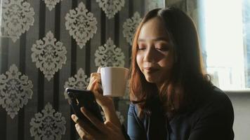 Mujer asiática bebiendo café mientras usa un teléfono celular para el trabajo