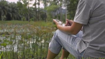 Woman sitting on the grass near natural pond using smartphone. video