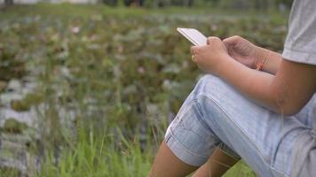 femme assise sur l'herbe près d'un étang naturel discutant sur smartphone. video