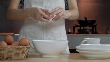 Female cook in a white apron is cracking an egg in home's kitchen.
