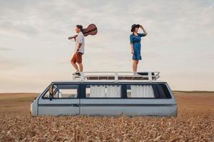 man with a guitar and woman are standing on roof of a car in a wheat photo