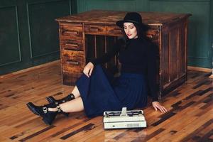 Beautiful woman in a hat sits near oak table with vintage typewriter photo