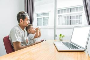 Padre e hijo asiáticos usando un teléfono inteligente y una computadora portátil juntos en casa foto