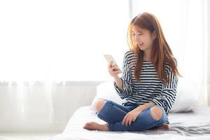 Young asian woman smiling sitting smart phone on bed.