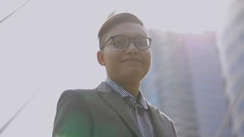 retrato bonito empresário asiático sorrindo e olhando para a câmera em pé na cidade urbana. homem asiático com óculos na rua perto de um grande prédio de escritórios. câmera lenta de baixo ângulo.