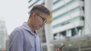Retrato de cámara lenta empresario asiático contesta el teléfono. Hombre asiático con gafas usa el teléfono en la calle cerca del gran edificio de oficinas. video