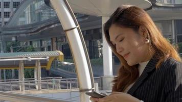 sorrindo, jovem mulher asiática, enviando mensagens de texto com um amigo no smartphone no fundo urbano da cidade, em busca de mídias sociais.