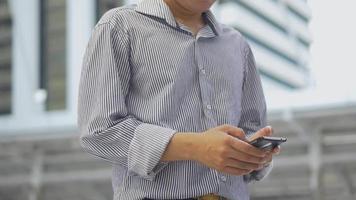 retrato empresário asiático em pé mensagens de texto com um amigo no smartphone no fundo urbano da cidade, em busca de mídias sociais. video