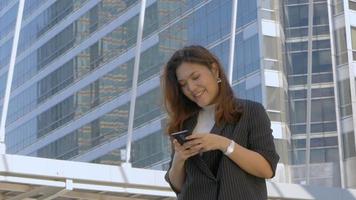sorrindo, jovem mulher asiática, enviando mensagens de texto com um amigo no smartphone no fundo urbano da cidade, em busca de mídias sociais. video