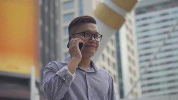 l'homme d'affaires asiatique de portrait au ralenti répond au téléphone. un homme asiatique avec des lunettes utilise un téléphone dans la rue près d'un grand immeuble de bureaux.