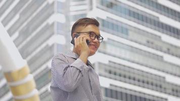 Retrato de cámara lenta empresario asiático contesta el teléfono. Hombre asiático con gafas usa el teléfono en la calle cerca del gran edificio de oficinas. video