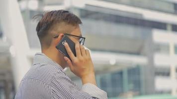 Retrato de cámara lenta empresario asiático contesta el teléfono. Hombre asiático con gafas usa el teléfono en la calle cerca del gran edificio de oficinas. video