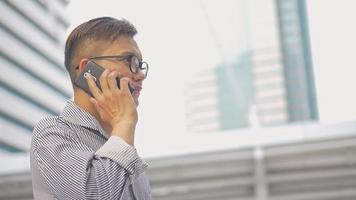 l'homme d'affaires asiatique de portrait au ralenti répond au téléphone. un homme asiatique avec des lunettes utilise un téléphone dans la rue près d'un grand immeuble de bureaux. video