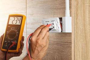 Electrician using a digital meter to measure the voltage at a wall socket on a wooden wall. photo