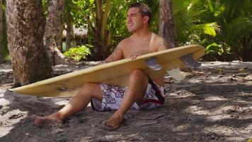 homem sentado na praia, encerando a prancha de surf, costa rica. filmado em vermelho épico para alta qualidade 4k, uhd, resolução ultra hd. video