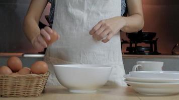 närbildsfilm framifrån, en kvinnlig kock i ett vitt förkläde knäcker ett ägg i en kopp för att förbereda en måltid på ett träbord i hemmets kök. äta äggulor är en hälsosam frukost. video