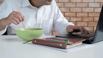 Thai male worker busy working with laptop, use chopsticks to hastily eat instant noodles during office lunch's break, because quick, tasty, and cheap. Over time Asian fast food, unhealthy lifestyle. video