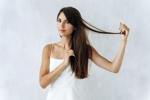 Cintura para arriba retrato de la mujer caucásica de pelo largo poniendo sus manos en su cabello y jugando con él mientras posa con una pared blanca en el fondo foto