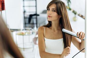 Beautiful young smiling woman using a hair straightener while looking into the mirror in bathroom. Confident girl looking at her reflection with pleasure smile photo