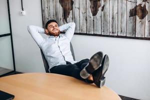 un chico atractivo con barba pone las manos detrás de la cabeza, pone los pies sobre la mesa y descansa. foto