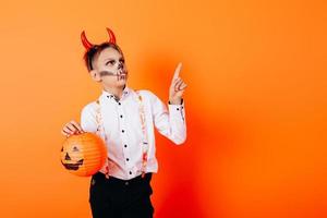 Boy in Devil masquerade makeup holding a pumpkin and pointing up. Halloween concept photo