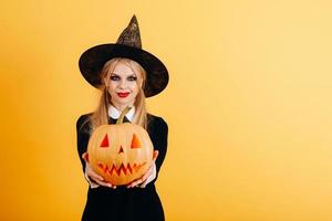 Mujer de pie sobre un fondo amarillo sosteniendo la calabaza y mostrándola con las manos rectas foto