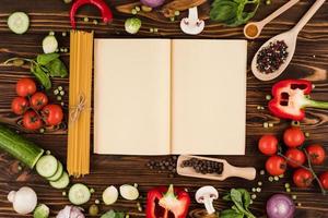 A recipe book is opened on a wooden table, with ingredients for Italian dishes laid out photo