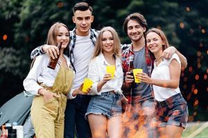 amigos en el campamento levantando una botella de cerveza y divirtiéndose mirando a la cámara foto