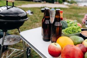 cerveza, salchichas y verduras frescas en la mesa al aire libre junto a la barbacoa foto