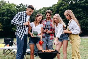 una compañía de amigos con una bebida y comida cocinando en la barbacoa del camping. - imagen foto