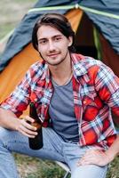 A guy sitting next the tent with  a bottle of beer and smiling looking at the camera photo