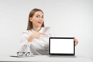 Smiling woman sits at the table and point to the screen  looking at the camera photo