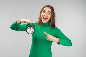 Laughing happy woman wearing casual clothes standing and holding alarm clock. Pointing to the clock photo