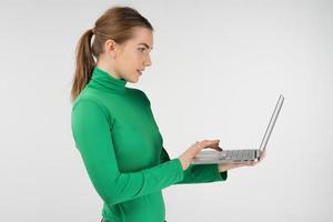 Mujer seria de perfil trabaja en un portátil sosteniéndolo en sus manos mientras está de pie contra el fondo blanco. foto