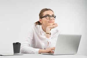 Sonriente mujer de negocios sentada detrás de una computadora portátil con una taza de café y un ipad en la mesa y mirando hacia arriba soñadora foto