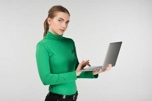 Woman in half-turn works on a laptop holding it in her hands  while standing against on the white background. Looks at the camera photo