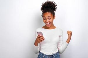 Feliz mujer mulata sosteniendo un teléfono inteligente en la mano y gritando con gesto ganador mirando la pantalla foto
