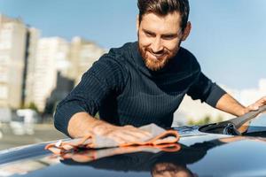 Cropped view of the bearded man with grey microfiber wipe polishing his new electric car. Handsome caucasian man washing and wiping his car with pleasure smile. Car washing concept photo