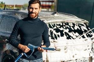 lavado de autos. Vista vertical de la cintura para arriba del hombre caucásico con equipo especial y sonriendo a la cámara. coche de limpieza utilizando el concepto de agua a alta presión. enfoque selectivo foto