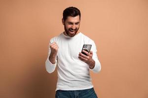 Feliz hombre lleno de alegría con barba mirando el teléfono inteligente y sonriendo haciendo un gesto de sí, celebrando la victoria o un buen trato. Foto de estudio de interior aislado sobre fondo beige